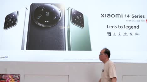 pedestrians walking past a commercial advertisement from the chinese tech brand xiaomi showcasing the xiaomi 14 series smartphone outside the official flagship store.