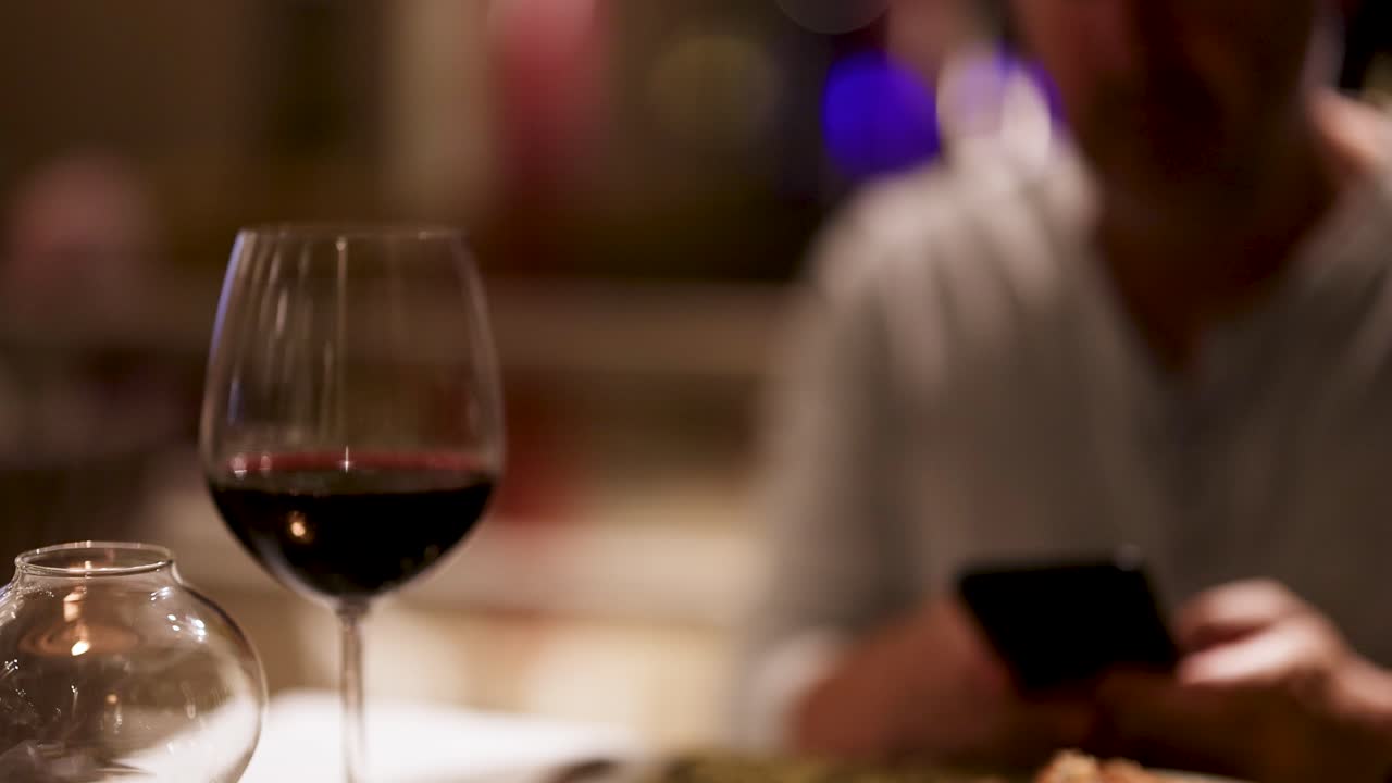A man uses his phone at a restaurant table with wine, creating a relaxed, intimate atmosphere with soft lighting