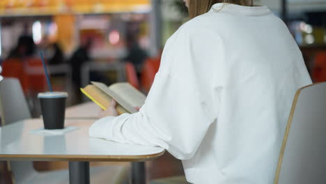 back view of young woman reading book at table in mall with coffee beside her, soft blur background featuring shoppers and people passing by