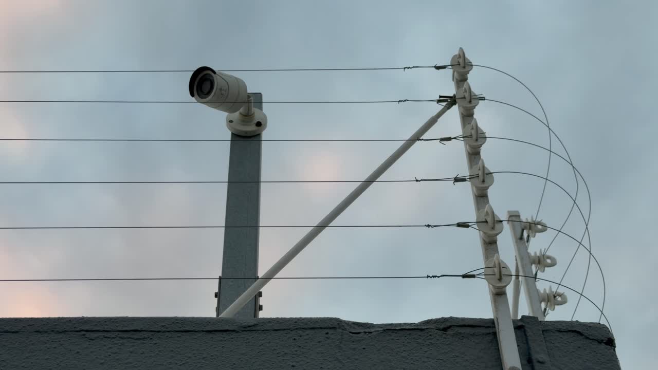 A security camera stands on a against a wall behind a electric fence