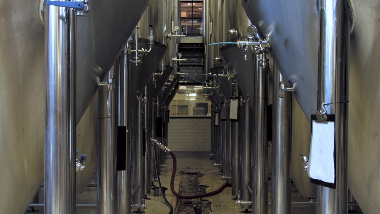 Slow tilt up from floor drain to rows of large stainless steel beer fermentation tanks in a brewery