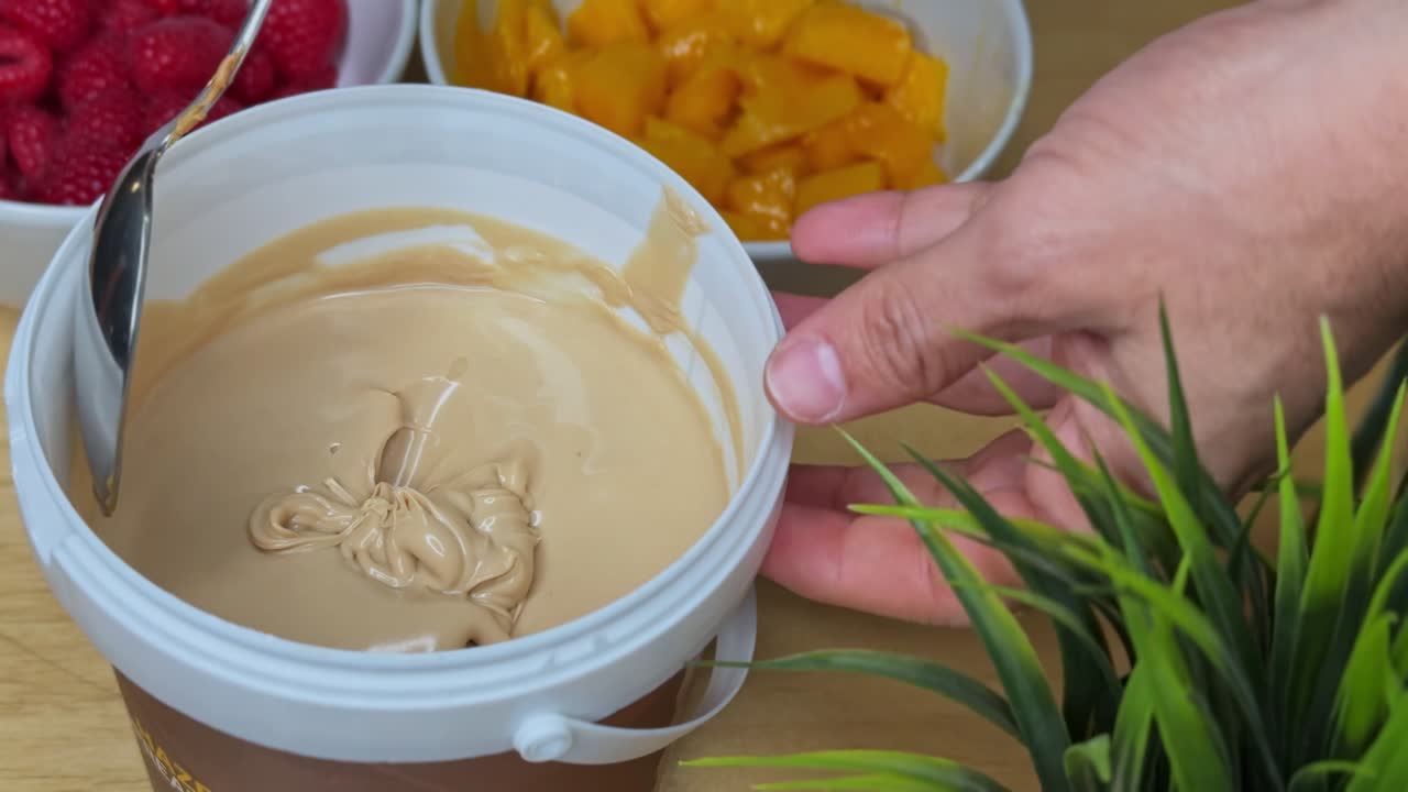 Close-up of a spoon scooping creamy caramel spread in a white tub with vibrant fruit in the background. Ideal for food content, recipe intros, product features, or kitchen-themed ads.