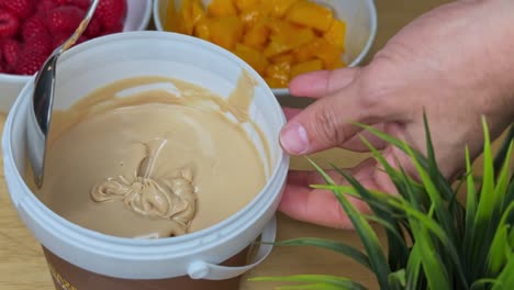 Close-up of a spoon scooping creamy caramel spread in a white tub with vibrant fruit in the background. Ideal for food content, recipe intros, product features, or kitchen-themed ads.