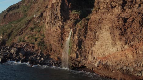 pequeña cascada hermosa que desemboca en el océano desde la pared de la montaña con olas que chocan contra ella durante la puesta de sol en ponta do sol, madeira, portugal