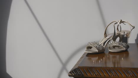 Slow-orbit-shot-of-elegant-diamond-bridal-shoes-sitting-on-mahogany-table-top-in-the-light-of-a-bay-window