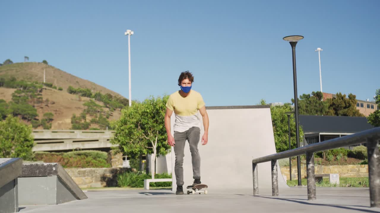 Caucasian man wearing face mask, riding and jumping on skateboard on sunny day