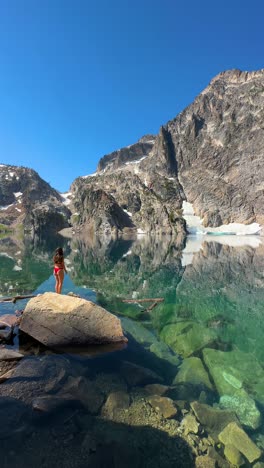 4k vertical, mujer joven en bikini de pie en la roca por encima de un hermoso lago alpino bajo colinas rocosas