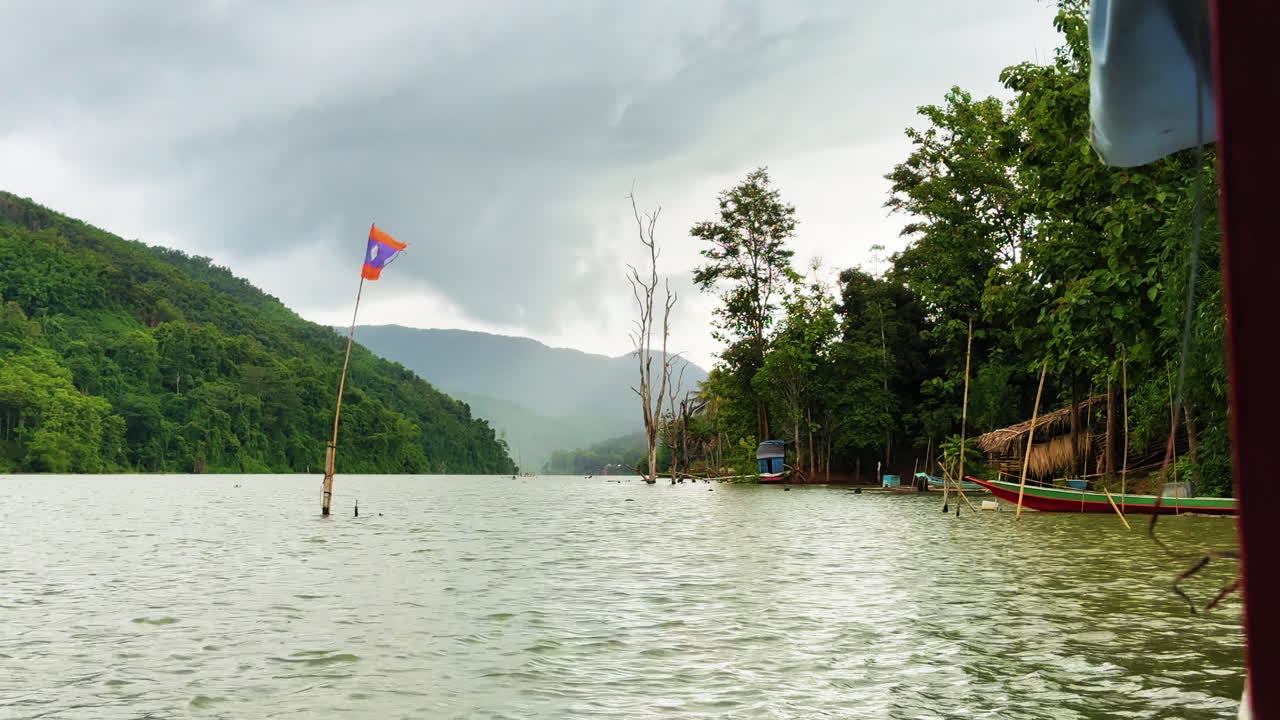 Il placido fiume Nam Ou nel nord del Laos con barche a coda lunga, sponde alberate e un segnale di bandiera, visto da una barca di passaggio sotto nuvole pesanti in una remota valle montana