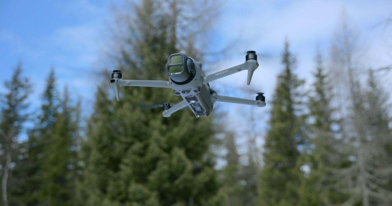 A Drone Hovers Above the Snow-covered Ground, Surrounded by a Forested Landscape in the Background - Tracking Shot
