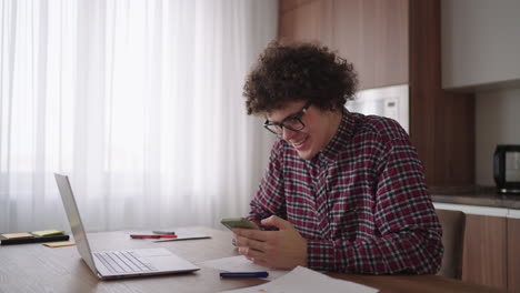 Estudiante-Masculino-De-Pelo-Rizado,-Un-Joven-Atractivo-Con-Gafas-Está-Estudiando-En-Casa-Usando-Una-Computadora-Portátil-Escribiendo-En-Un-Cuaderno.-Estudiante-Universitario-Usando-Una-Computadora-Portátil-Viendo-Un-Seminario-De-Aprendizaje-En-Línea-A-Distancia