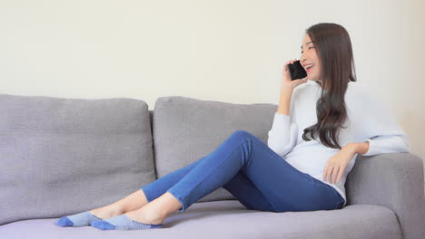 Asian-Brunette-Charming-Woman-Sitting-on-the-Gray-Sofa-and-Talking-on-the-Phone-Joyfully