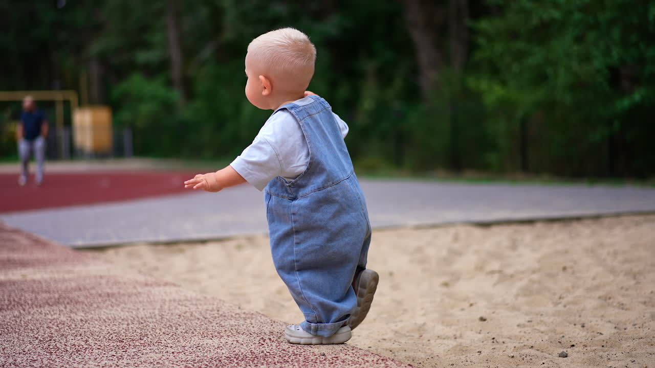 Little cute kid in a loose jeans romper walks up to a ball. Lovely toddler plays on the playground outdoors in summer.