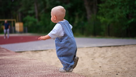 little cute kid in a loose jeans romper walks up to a ball. lovely toddler plays on the playground outdoors in summer.