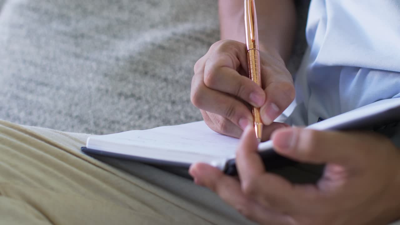 Adult Asian man sitting on couch holding smartphone, stabilizing notebook, writing with gold pen
