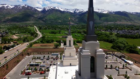 drone flying up while looking at layton lds mormon temple steeple reviling golden angel moroni with beautiful wasatch mountain range in background with blue sky and fluffy white clouds