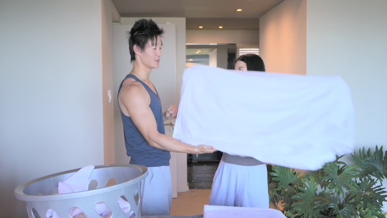 Asian couple starting with plastic laundry basket on bed, folding towels, organizing linen, hugging