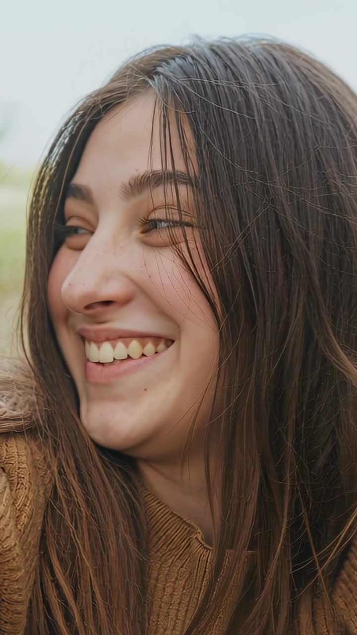 Video vertical: Adolescente riendo e inclinándose hacia la cámara, el obturador de la cámara haciendo clic, en un parque, llevando un suéter marrón.