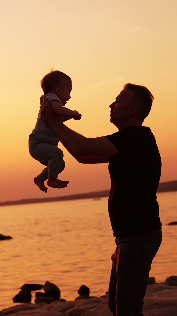 Father and baby boy spending time at the river bank. Dad tossing up his little son at backdrop of orange sky at sunset. Vertical video