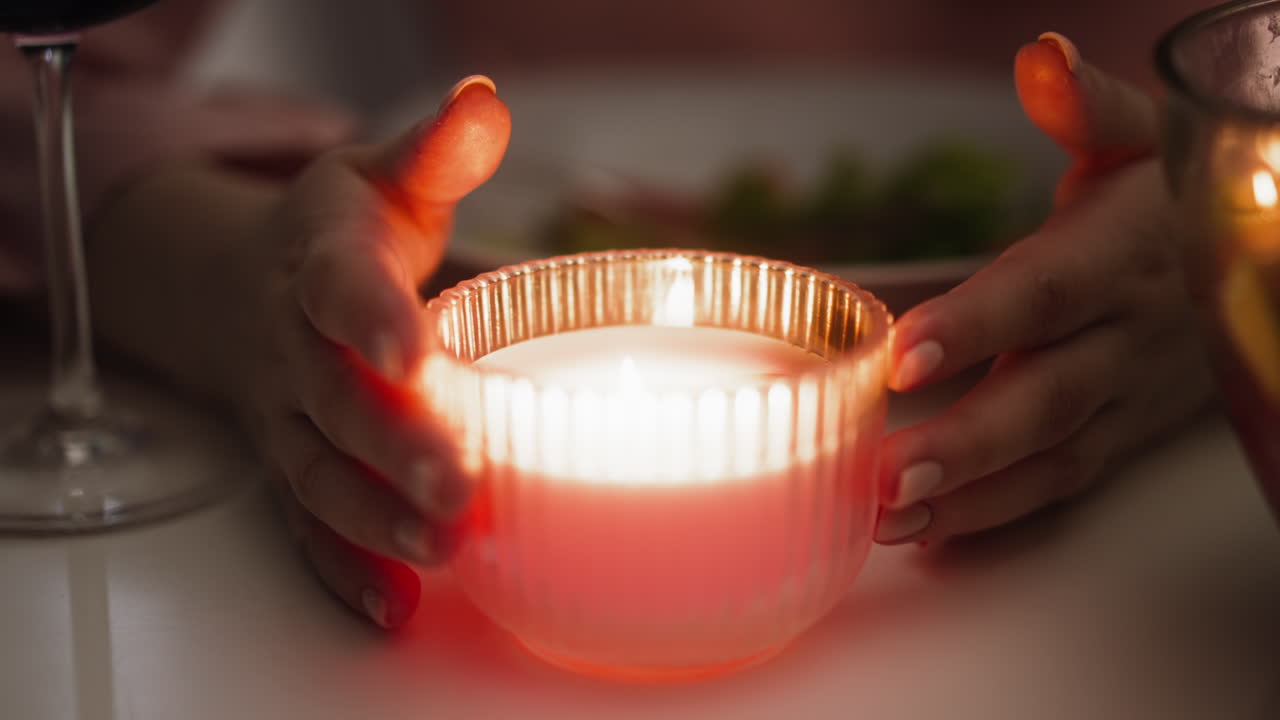 женщина поворачивает горящую свечу в стеклянном держателе, сидя за столом