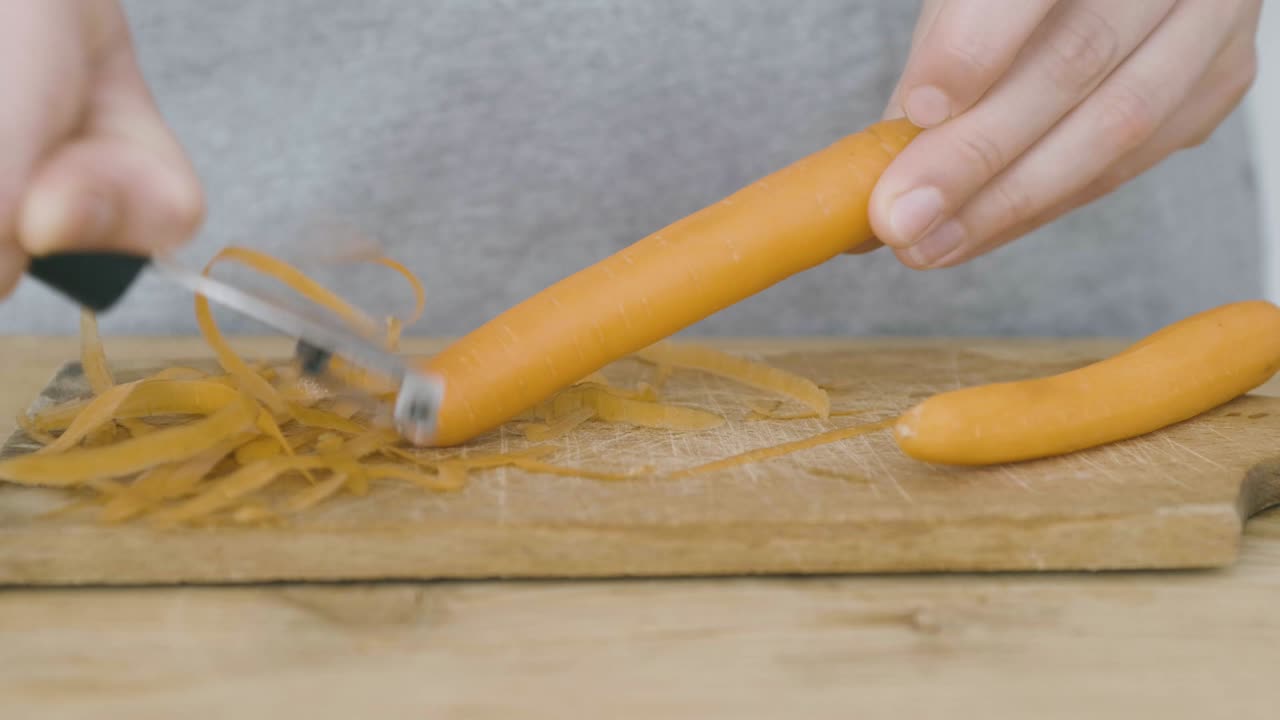 Two hands peel a carrot using a potato peeler on a wooden cutting board, a carrot has already been peeled
