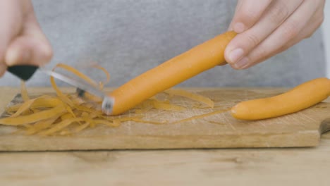 two hands peel a carrot using a potato peeler on a wooden cutting board, a carrot has already been peeled