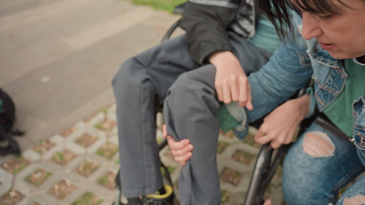 White Woman Adjusting Wheelchair Shoe Outdoors, Denim Jacket Reaches To Fasten Strap For Seated Boy, Sidewalk Park Setting, Close Interaction Showing Empathy, Gentle Kneeling Motion And HandsOn
