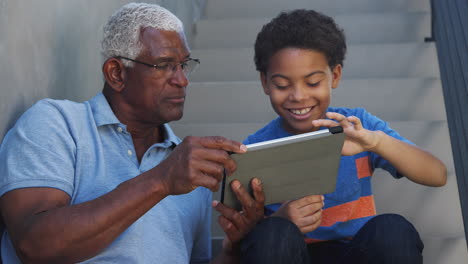 grandfather with grandson sitting on steps outdoors at home using digital tablet