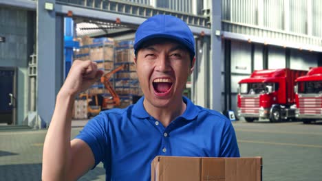 close up of asian male courier in blue uniform celebrating succeed delivering a carton, outside of logistics distributions warehouse