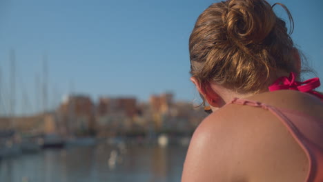 Woman-reading-something-wearing-sunglasses-on-ocean-coastline-with-blurred-boats,-back-view