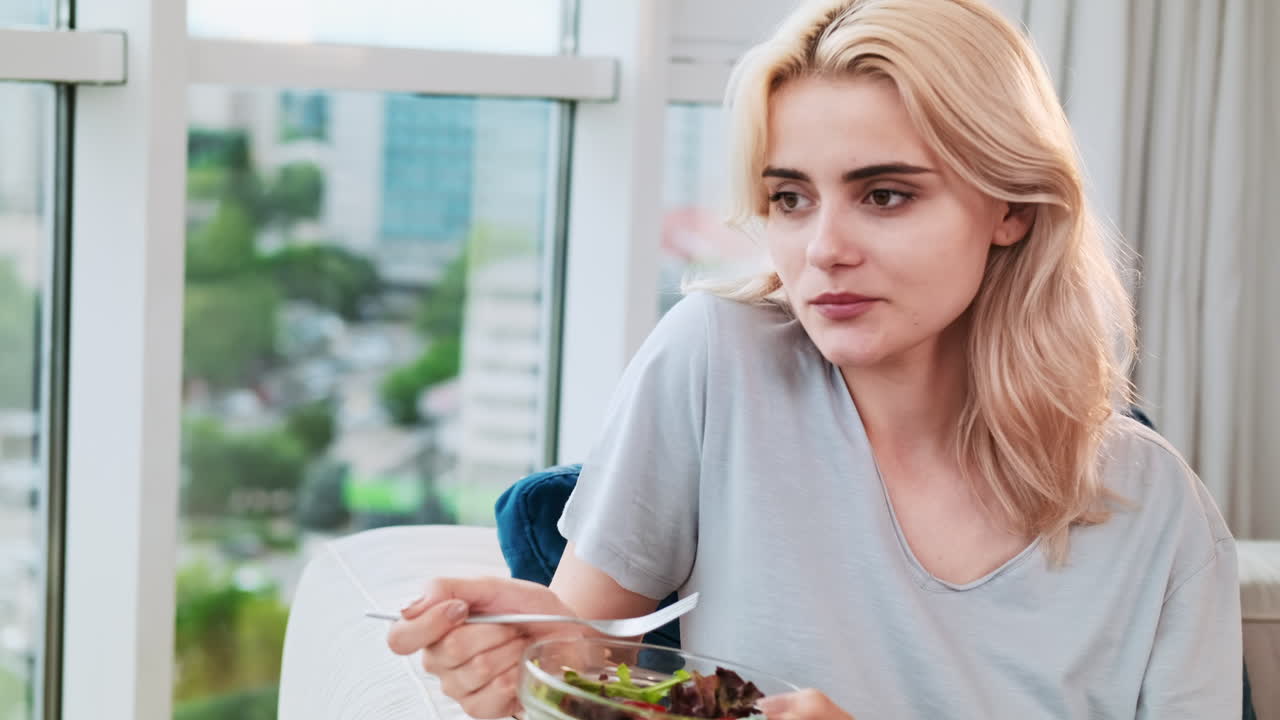 Young blonde smiling woman sitting near the panoramic window and eating healthy salad