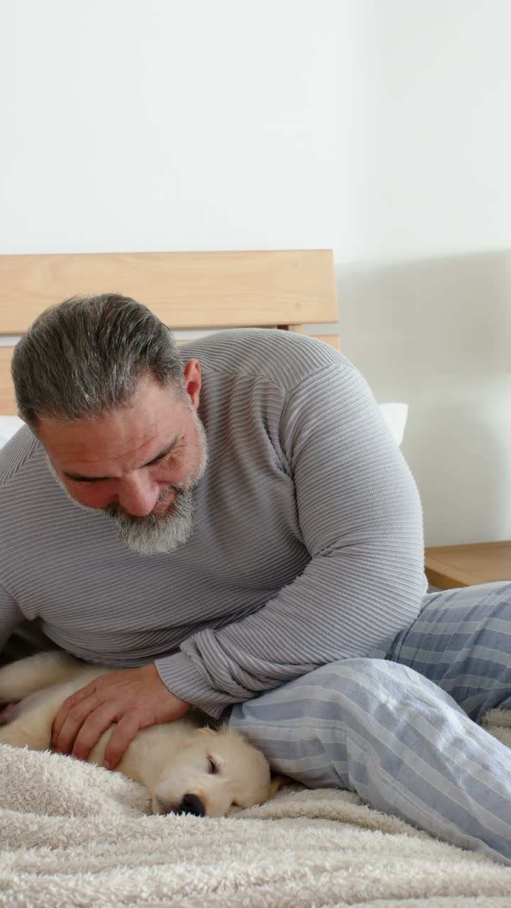 Video verticale: uomo bianco appoggiato alla testiera accarezza un cucciolo su una coperta in camera da letto, calmandolo