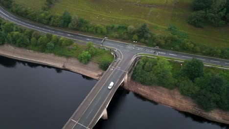 aerial view of white van crossing bridge over calm reservoir, surrounded by lush green countryside