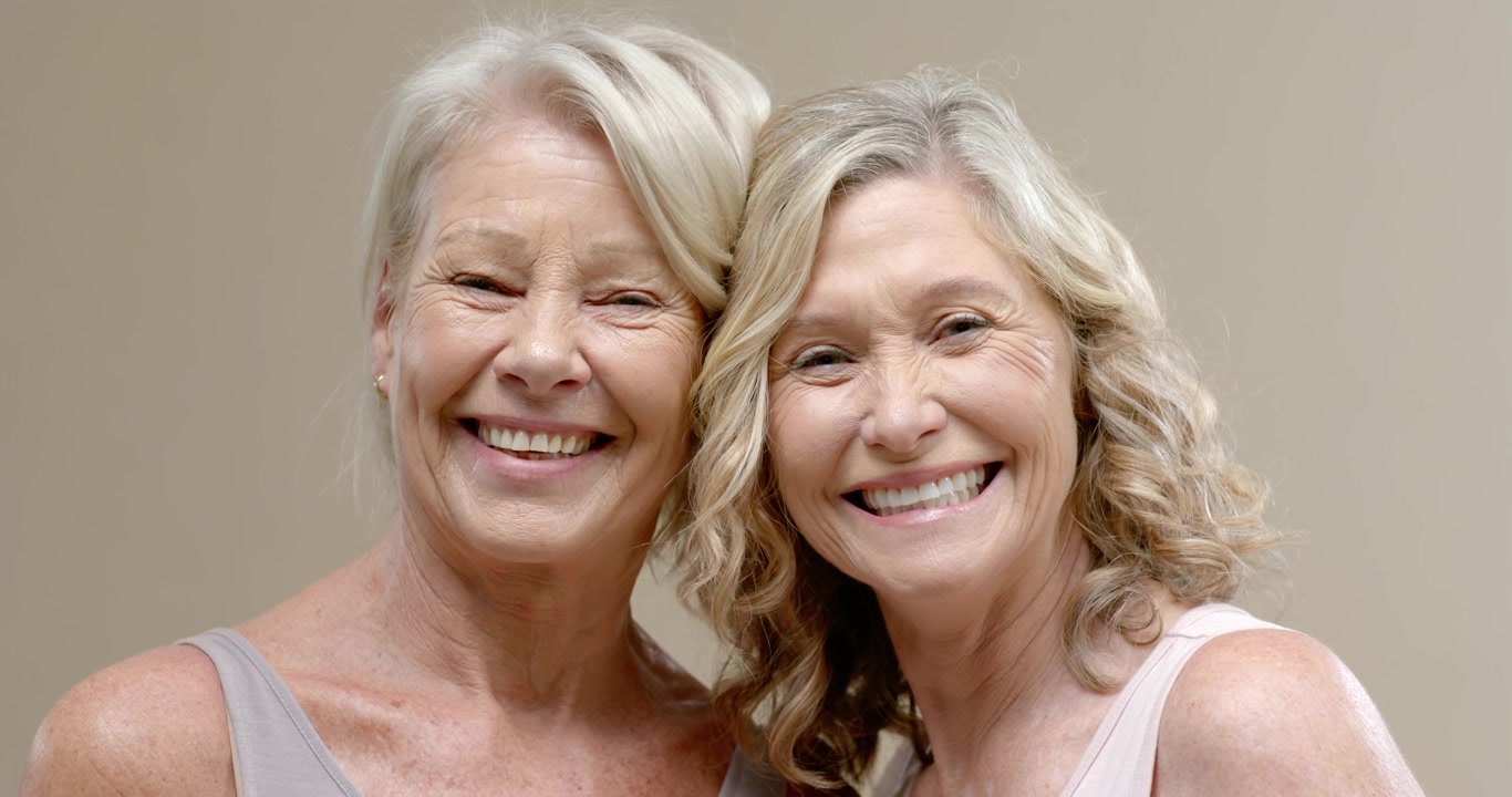 Senior women sharing amused glance, smiling and leaning closer in beige studio with tank tops
