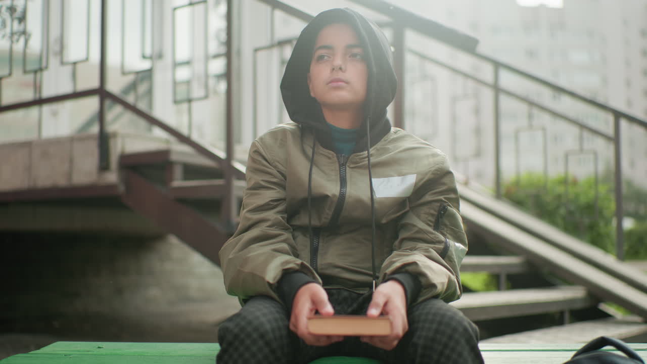 Reflective little boy in hooded jacket seated outdoors holding book on lap, gazing thoughtfully into distance with partial view of backpack by the side near staircase background in urban environment