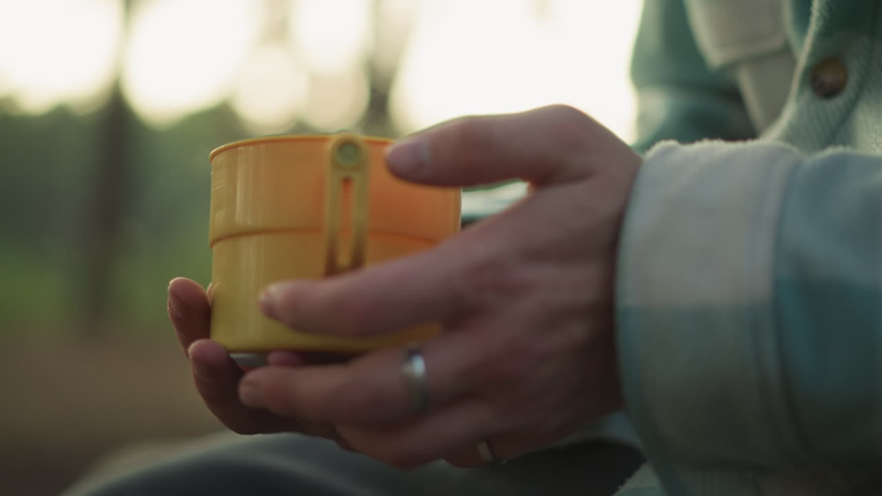 Persona che stringe una tazza nella soffice luce della foresta, scena intima di mani che tengono una bevanda calda in un ambiente tranquillo, primo piano di una persona che tiene delicatamente una tazza in mezzo al bagliore dorato e alla tranquillità della foresta