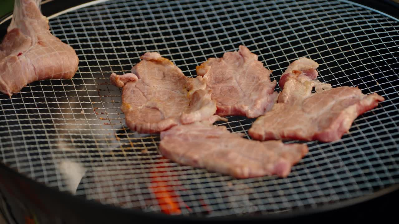 A person uses metal tongs to flip sizzling slices of fresh pork, revealing the cooked side while grilling over a hot charcoal fire during a summer barbecue