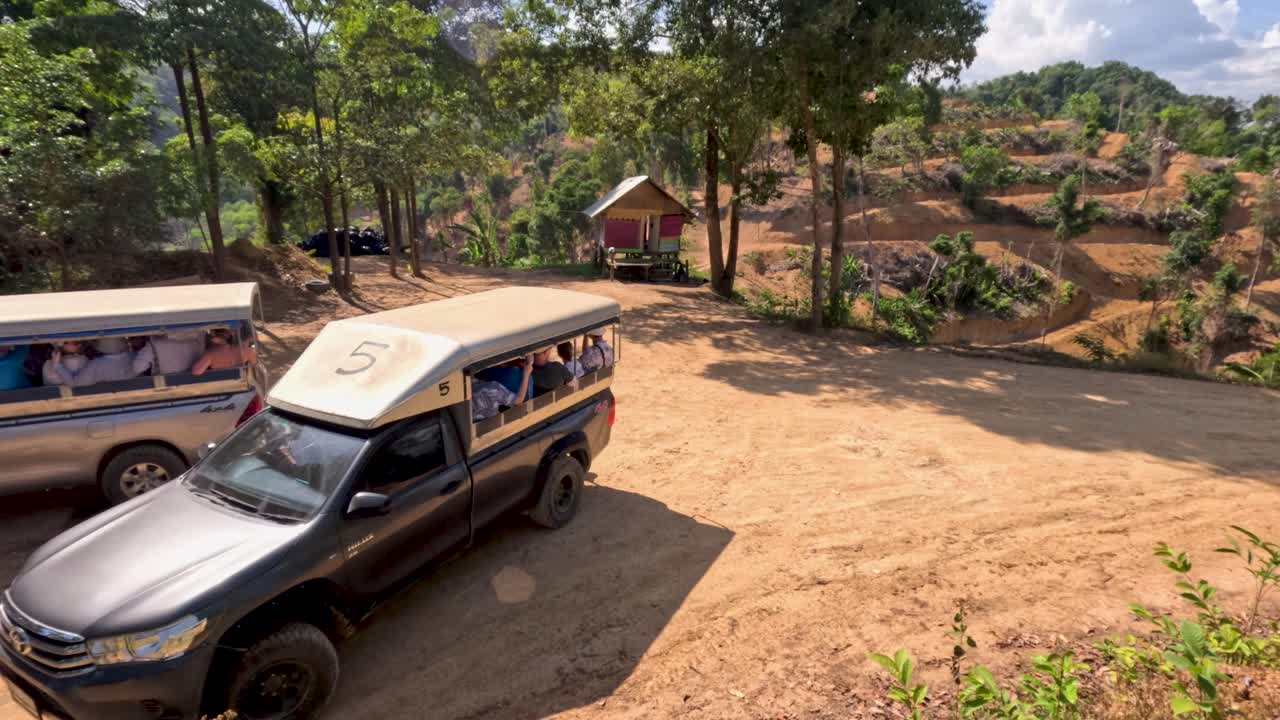 Tourist trucks arrive on dirt road in sunny jungle, passengers observing, wide shot, natural lighting
