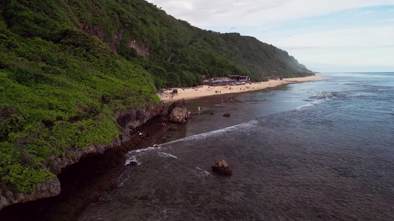 Drone video reveals a calm tropical beach along a wide Bali shoreline where low tide exposes shallow reefs beside a steep coastal green hill dotted with visitors enjoying the peaceful ocean atmosphere