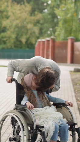 redhead woman leans to kiss little girl with disability sitting in wheelchair. mother enjoys spending time with daughter in city park on spring day