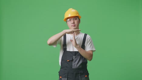 asian man worker wearing goggles and safety helmet looking at camera and showing time out hands gesture while standing in the green screen background studio