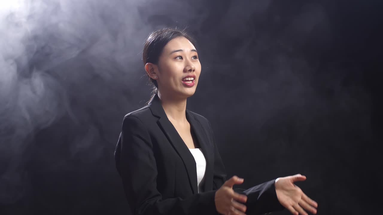 vista lateral de la oradora asiática mujer en traje de negocios mostrando los dedos índice hacia arriba mientras habla en el estudio de pantalla negra