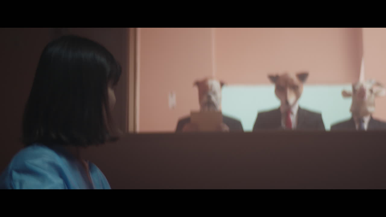 A girl sits at a table and strange figures in animal masks observes her from behind the glass