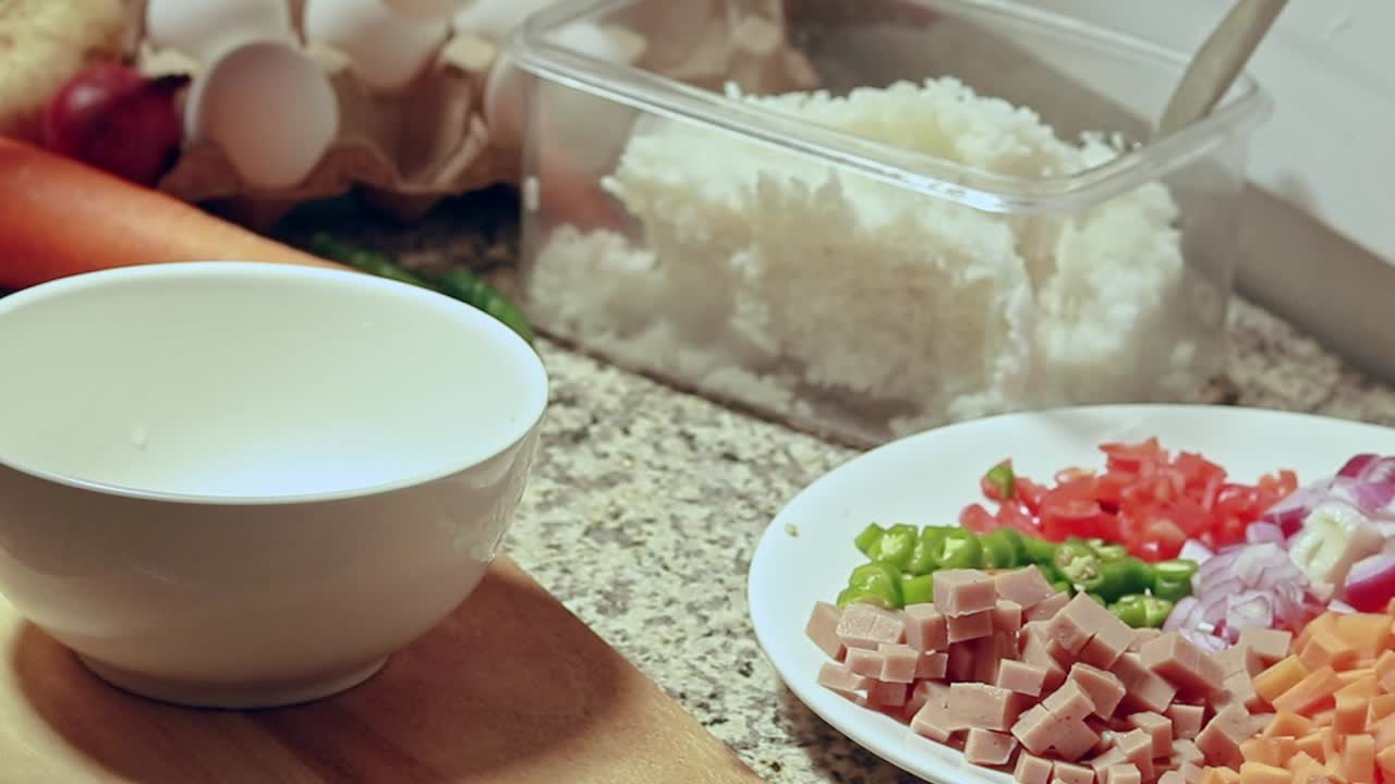 Setting a white bowl on a kitchen counter beside a plate of diced vegetables and ham and cracking an egg on it. Showing cooking fried rice at home