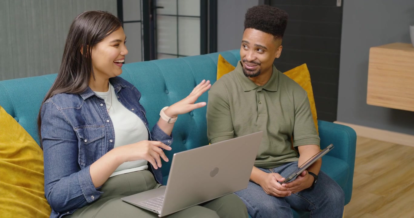 Pareja afroamericana sentada en un sofá color verde azulado, la mujer gesticulando, revisando el contenido de una laptop y una tablet.