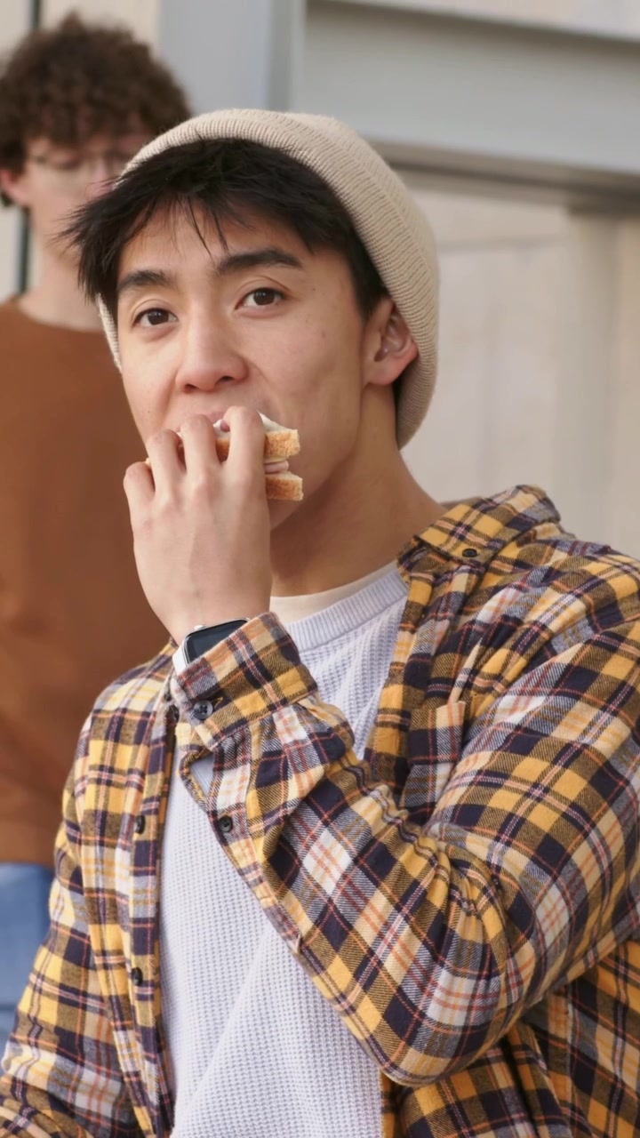 縦型動画：ビーニー帽をかぶったアジア人男性が、昼食にファサードの前でサンドイッチをかじりながら、左をちらりと見ている様子。