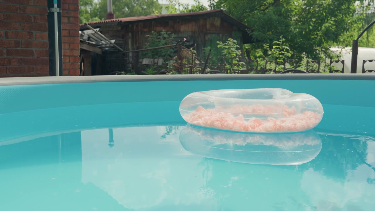Clear plastic pool float filled with pink salt rests on calm blue water in backyard setting. weathered brick wall and green foliage are visible beyond edge of swimming pool creating tranquil