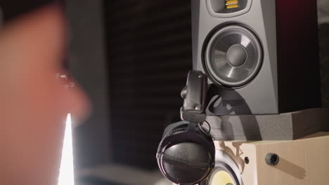 close-up of a blurred face in the foreground, with a recording studio setup visible in the background, including a speaker and a pair of headphones hanging on it