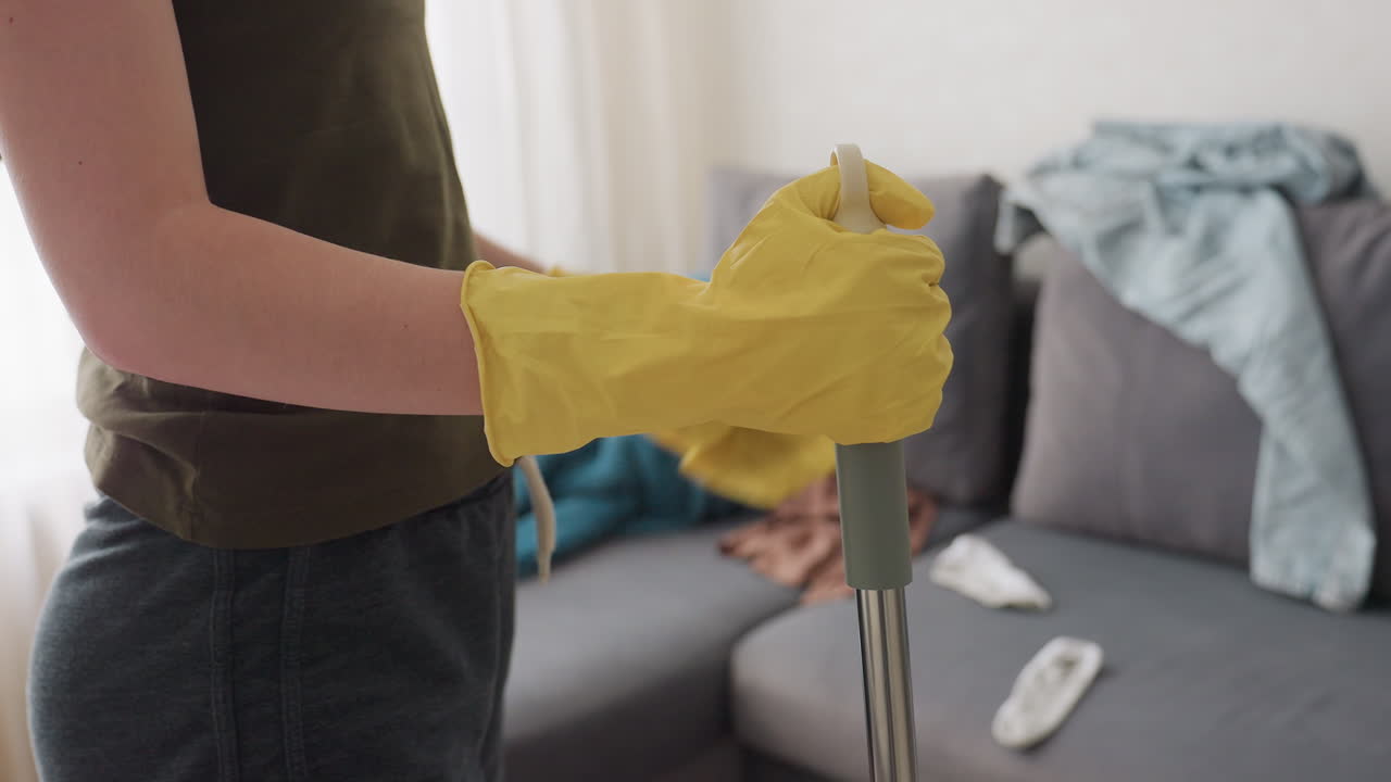 Close up side view of woman in yellow gloves holding mop handle while standing near couch with scattered clothes and blanket in bright room interior showing daily cleaning routine