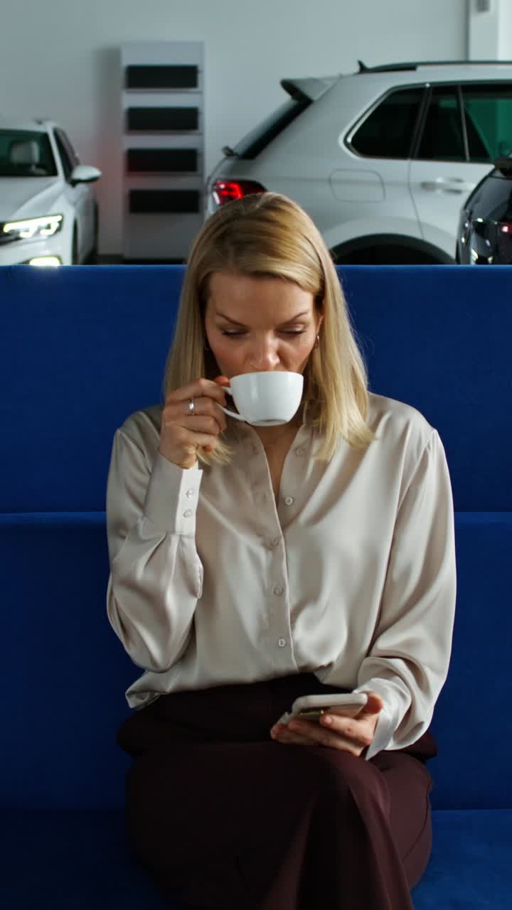 여성이 커피를 마시며 자동차 전시장에서 전화를 사용하고 있습니다.
