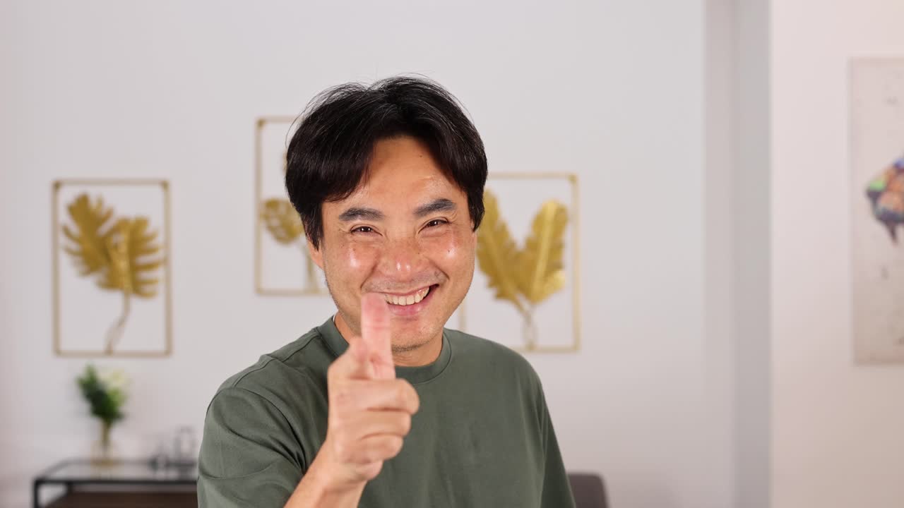 Middle-aged Asian man smiles and points at the camera with a friendly gesture in a well-lit, modern living room with decorative wall art